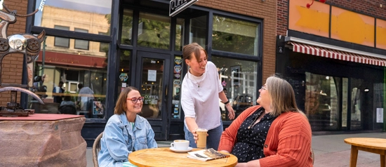 A waitress smiling while placing an iced coffee on a table outside where two women are seated